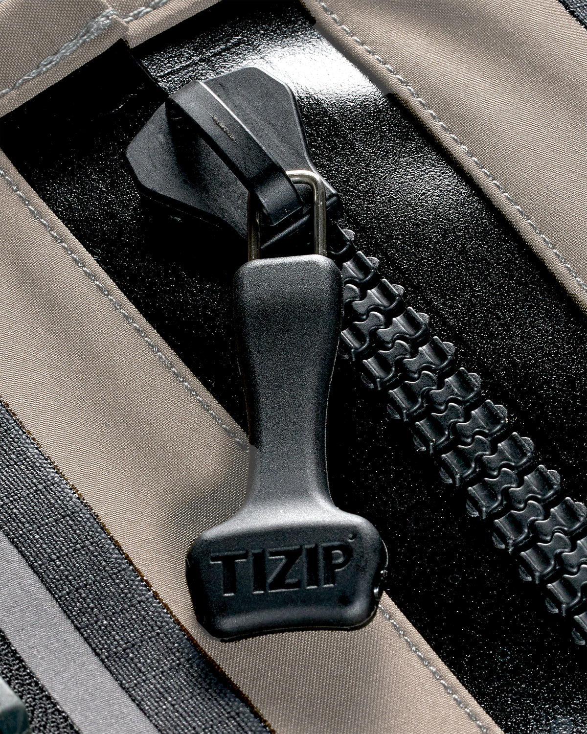 Close-up of a black TIZIP zipper pull on a beige and black fabric bag, showing the brand name embossed on the pull.